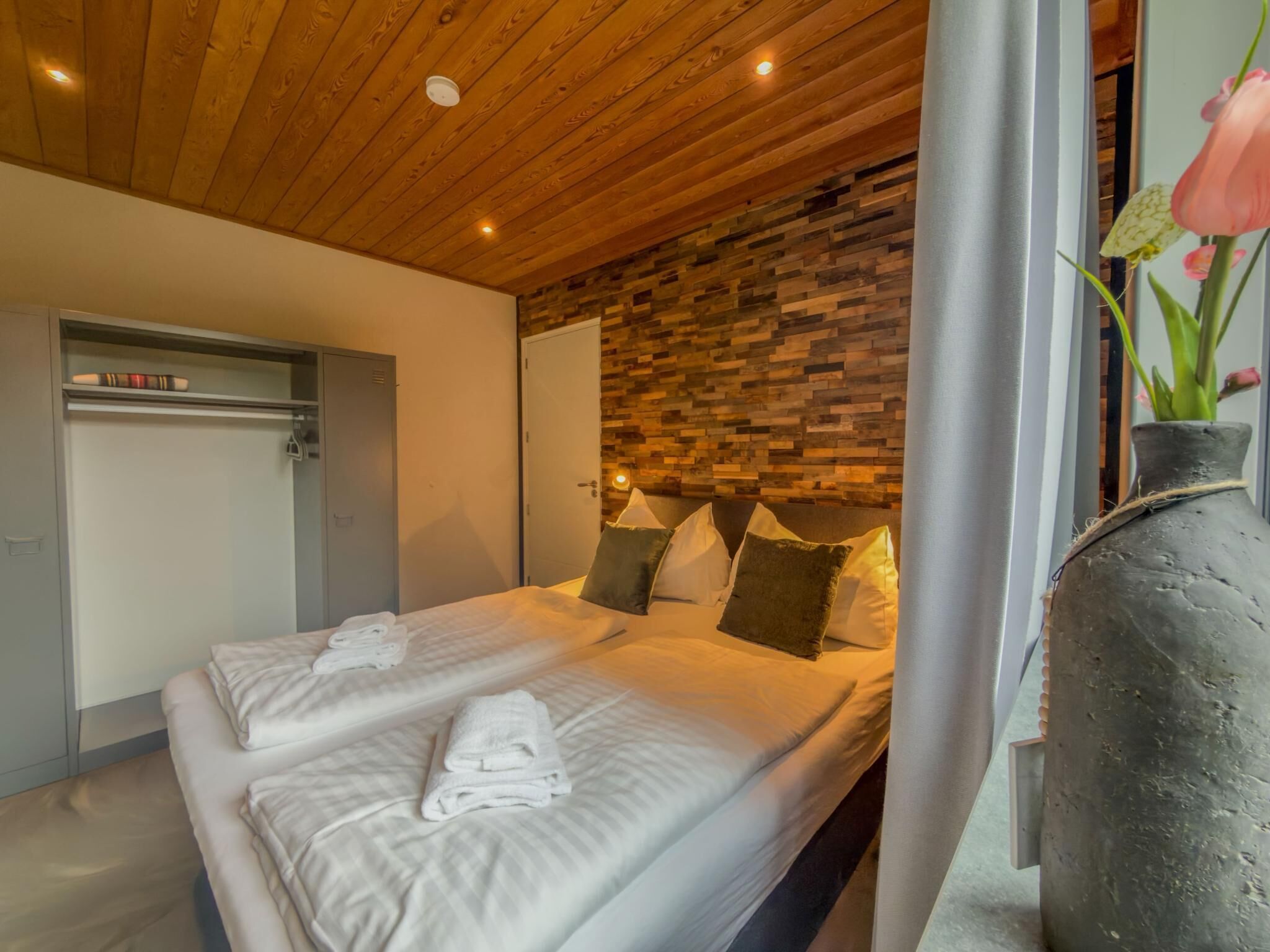A hotel room with a wooden ceiling, a bed with white linens, a stone wall, and a vase with flowers.