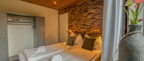 A hotel room with a wooden ceiling, a bed with white linens, a stone wall, and a vase with flowers.