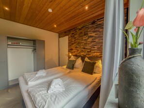 A hotel room with a wooden ceiling, a bed with white linens, a stone wall, and a vase with flowers.