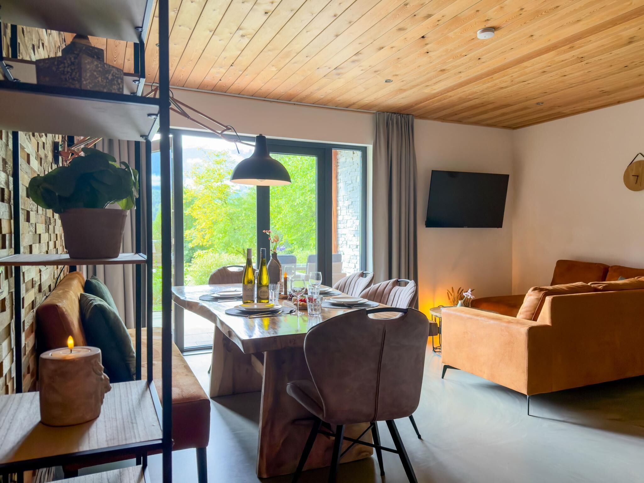 A modern living room with a dining area, wooden ceiling, large windows, and a flat-screen TV.