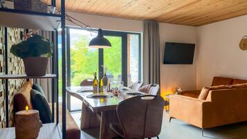 A modern living room with a dining area, wooden ceiling, large windows, and a flat-screen TV.