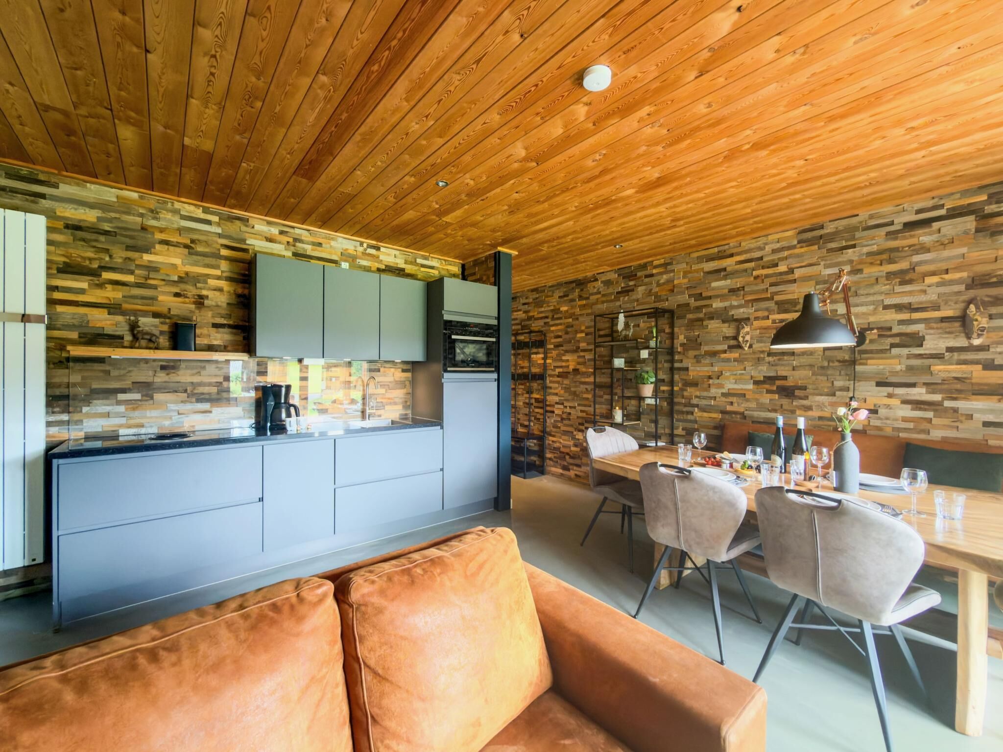 A modern kitchen with wooden cabinets and a stone backsplash, a dining area with a wooden table and chairs, and a leather sofa.