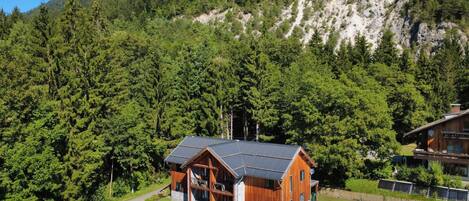 A wooden cabin with a metal roof, surrounded by dense forest and mountains.