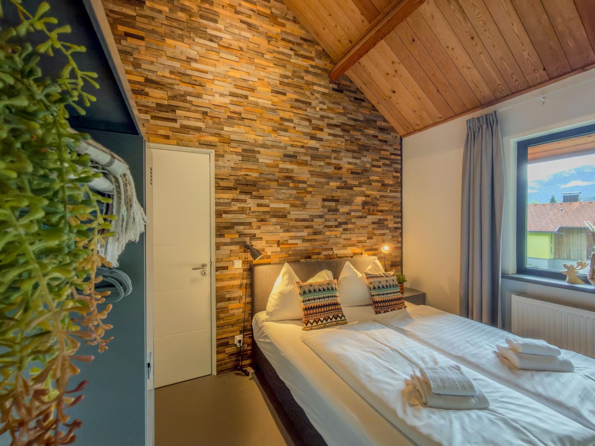 A bedroom with a wooden ceiling, a brick-patterned wall, a bed with white linens, and a window with a view of buildings.