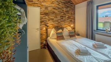 A bedroom with a wooden ceiling, a brick-patterned wall, a bed with white linens, and a window with a view of buildings.