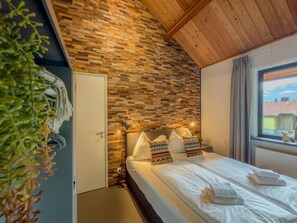 Un dormitorio con techo de madera, pared con estampado de ladrillo, cama con ropa blanca y ventana con vista a edificios.