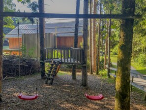 Un'area giochi con altalene in legno e uno scivolo, circondata da alberi e un edificio con tetto in metallo.