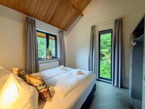 Ein Schlafzimmer mit einem Bett, zwei Fenstern, einer Decke mit Holzbalken und Blick ins Grüne.