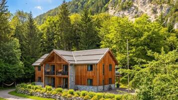 A two-story wooden cabin with a stone foundation, surrounded by lush greenery and a mountain backdrop.