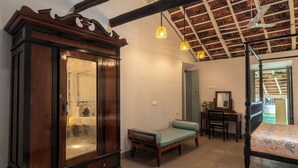 A room with a large wooden wardrobe, a bed with a floral bedspread, a bench, a desk with a chair, and a ceiling fan. - Casa Particular in Cansaulim, India (Arossim)