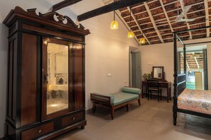 Ein Zimmer mit großem Holzschrank, einem Bett mit blumigem Überwurf, einer Bank, einem Schreibtisch mit Stuhl und einem Deckenventilator.