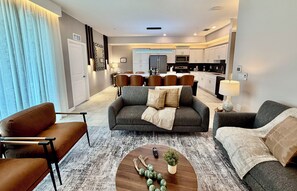 A modern living room with a sofa, armchairs, and a coffee table. There is a kitchen in the background with white cabinets and appliances. - 401 - Beyond the Sea & Into the Stars (Kissimmee)