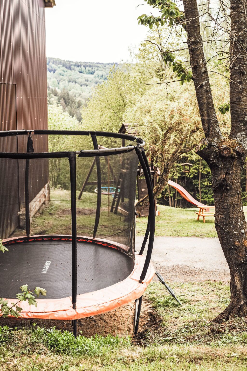 Ein Trampolin mit Sicherheitsnetz, umgeben von Bäumen und einer hölzernen Bank in einem Garten.