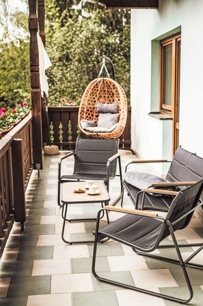 Un balcon avec deux fauteuils de relaxation noirs, un fauteuil suspendu en osier et une petite table sur laquelle se trouvent une tasse et des biscuits.