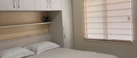 Un dormitorio con cama, armarios blancos, ventana con persianas y un equipo de aire acondicionado.