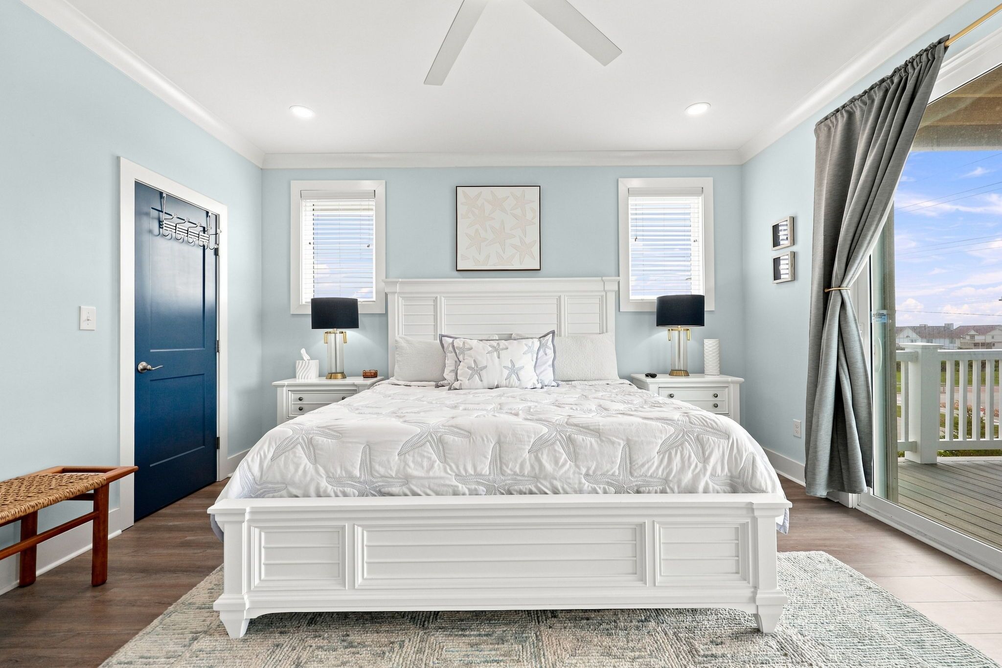 A bedroom with a large bed, two bedside tables, a window with blinds, a ceiling fan, and a view of the outside.