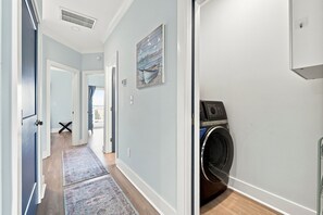 A laundry room with a front-loading washer and dryer, a hallway with a rug, and a door leading to another room. - Shore Perfection | Family Beach Retreat, Sleeps 8 (Emerald Isle)