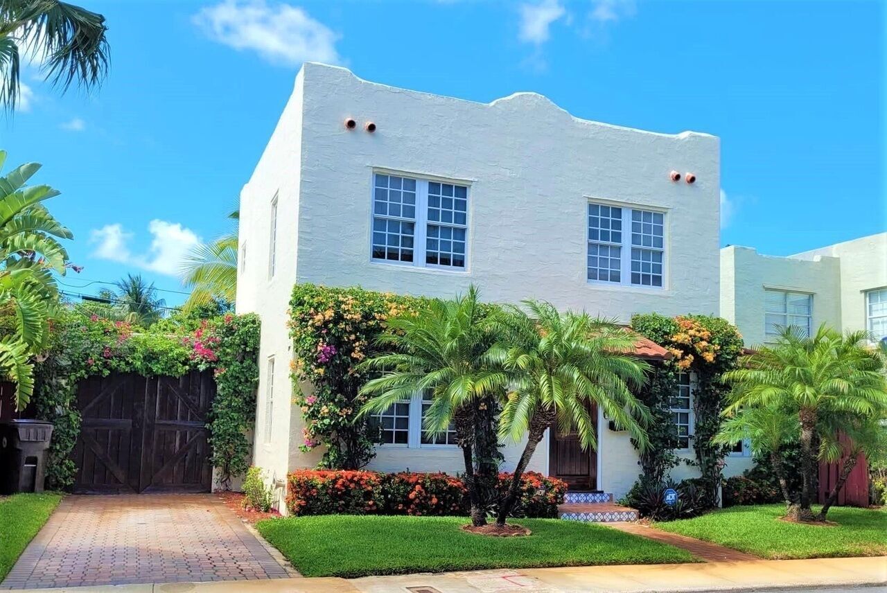 Un bâtiment à deux étages, à l’extérieur blanc, avec une porte d’entrée et un petit jardin devant, agrémenté de palmiers et de plantes à fleurs.