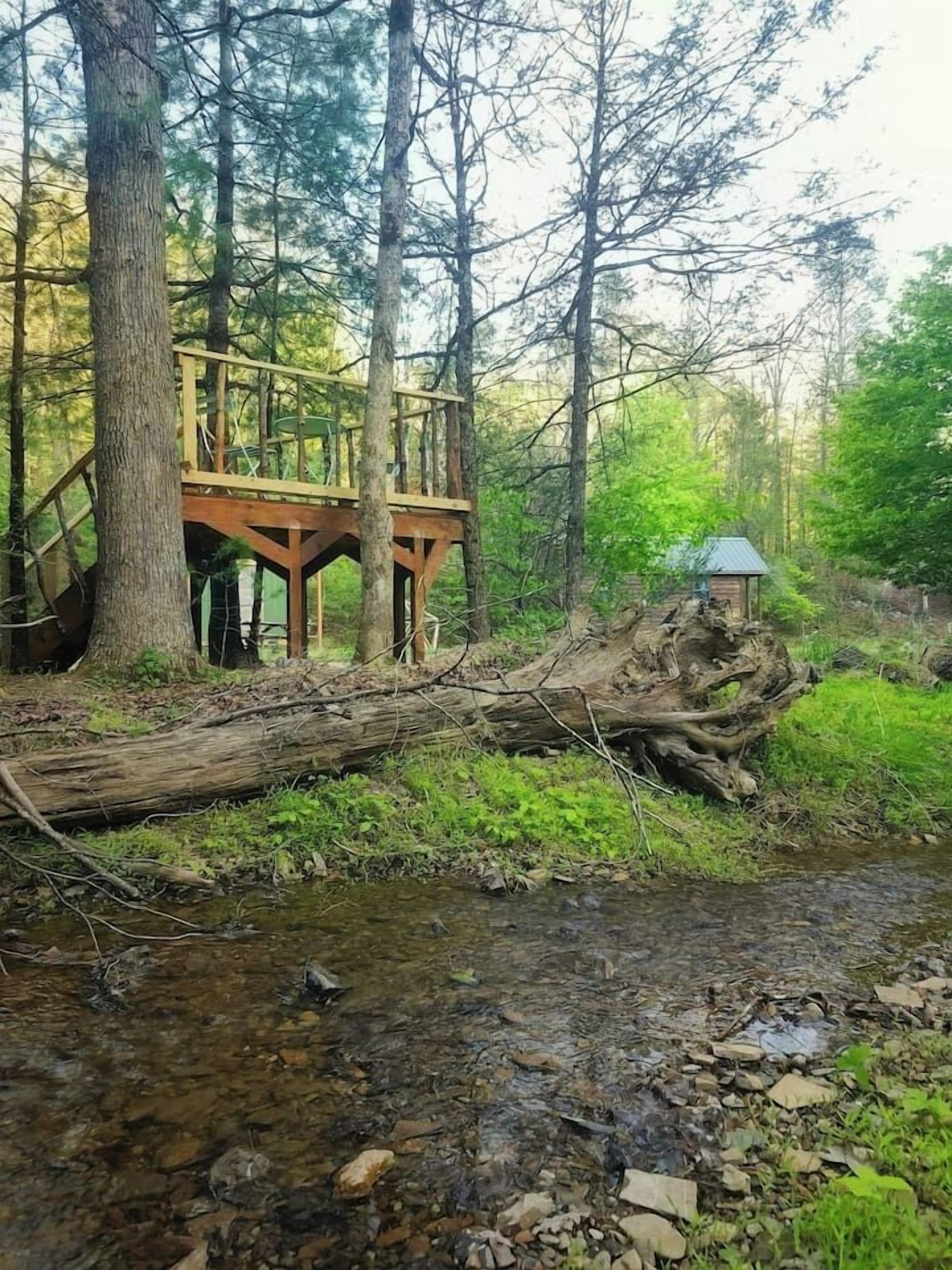 A wooden bridge over a stream with a rustic cabin and a gazebo in a forested area.