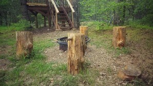 A forested area with a wooden staircase leading to a platform, surrounded by stumps and a fire pit.