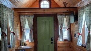 A cabin interior with a green door, wooden walls, a sloped ceiling, and two windows with curtains.