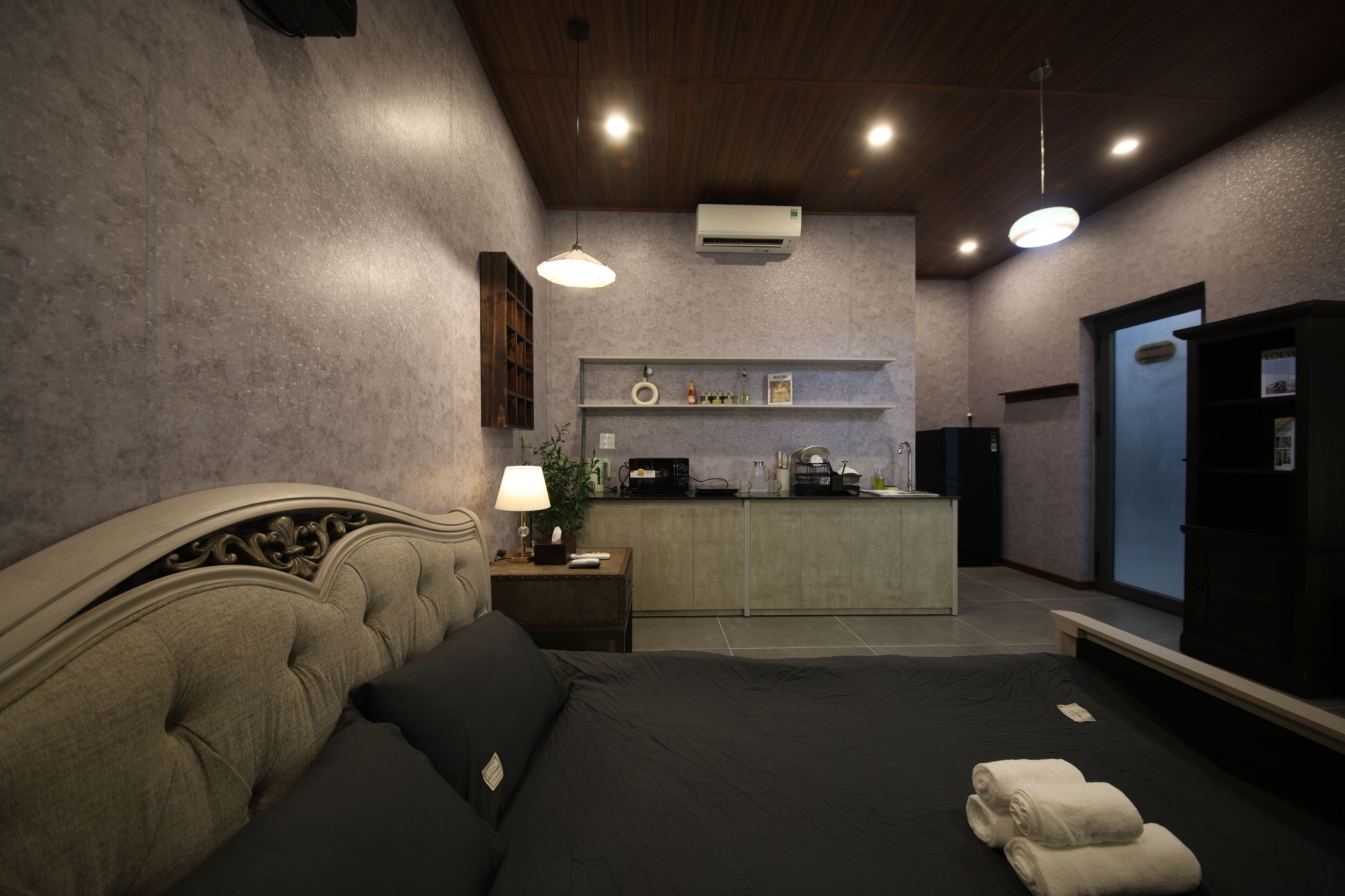 Quarto com cama, mesa de cabeceira, luminária e pequena área de cozinha.