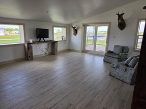 Ein geräumiges Wohnzimmer mit Holzboden, einer Wandhalterung für den Fernseher und einem großen Fenster mit Blick auf eine grüne Landschaft.