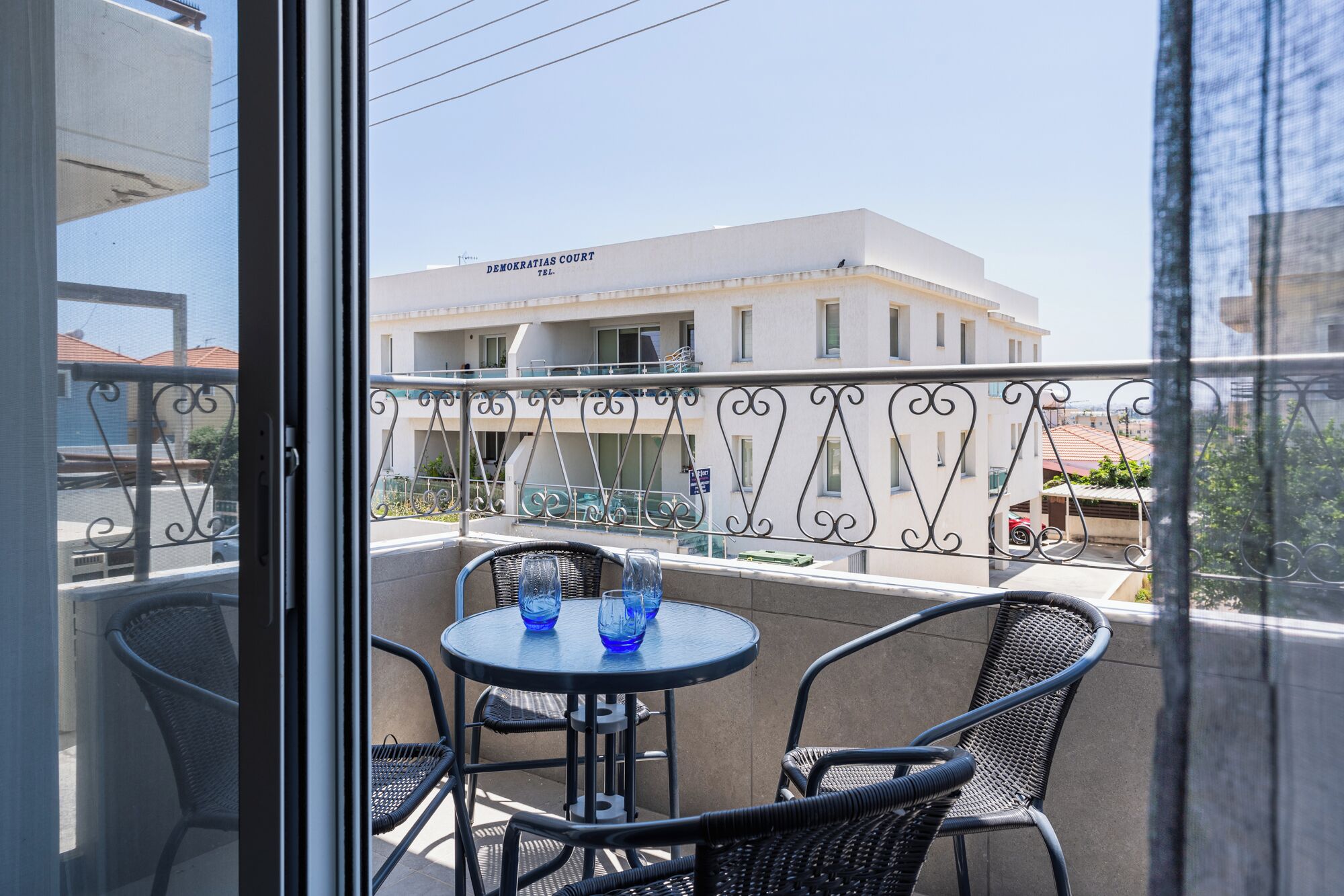 A balcony with a table and chairs, overlooking a building with a sign that reads 'Demokratias Court Tel.'
