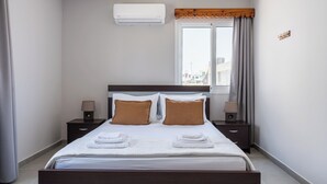 Hotellihuone, jossa on sänky, kaksi yöpöytää, ikkuna verhoilla, ilmastointilaite ja näkymä ulkona oleviin rakennuksiin.