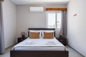 A hotel room with a bed, two bedside tables, a window with curtains, an air conditioning unit, and a view of buildings outside. - Modern 2BR Apt in Oroklini Larnaca (Voroklini)