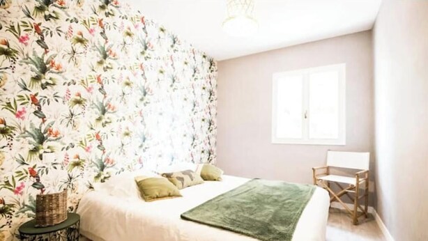 Una camera da letto con carta da parati floreale, un letto con cuscini, una coperta verde, un tavolino rotondo e una sedia di legno.