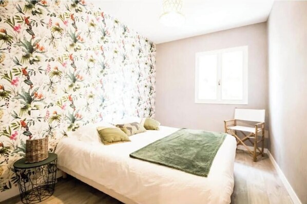 A bedroom with a floral wallpaper, a bed with pillows, a green throw, a small round table, and a wooden chair.