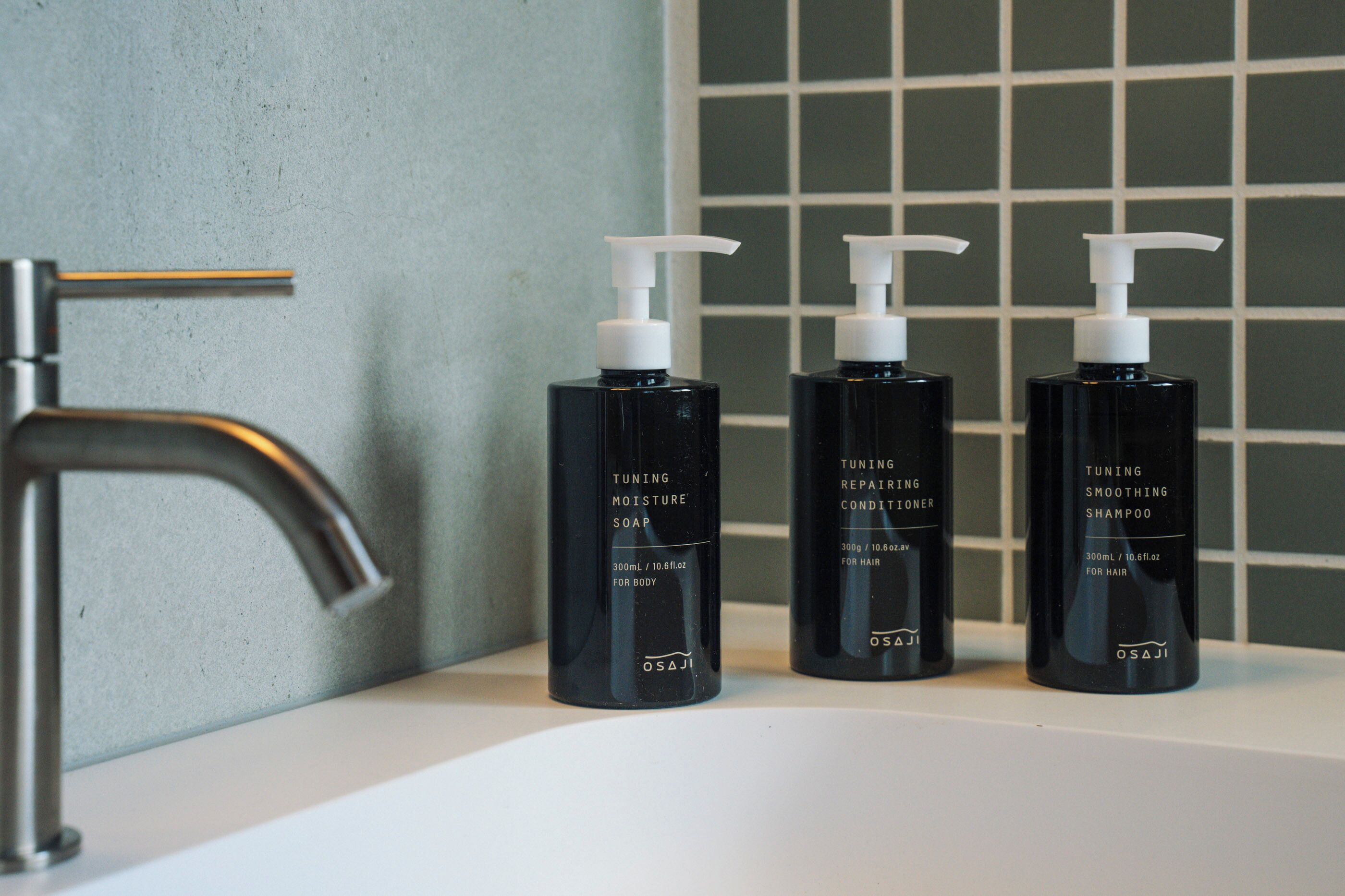Three black bottles of hair care products on a white surface against a tiled wall.