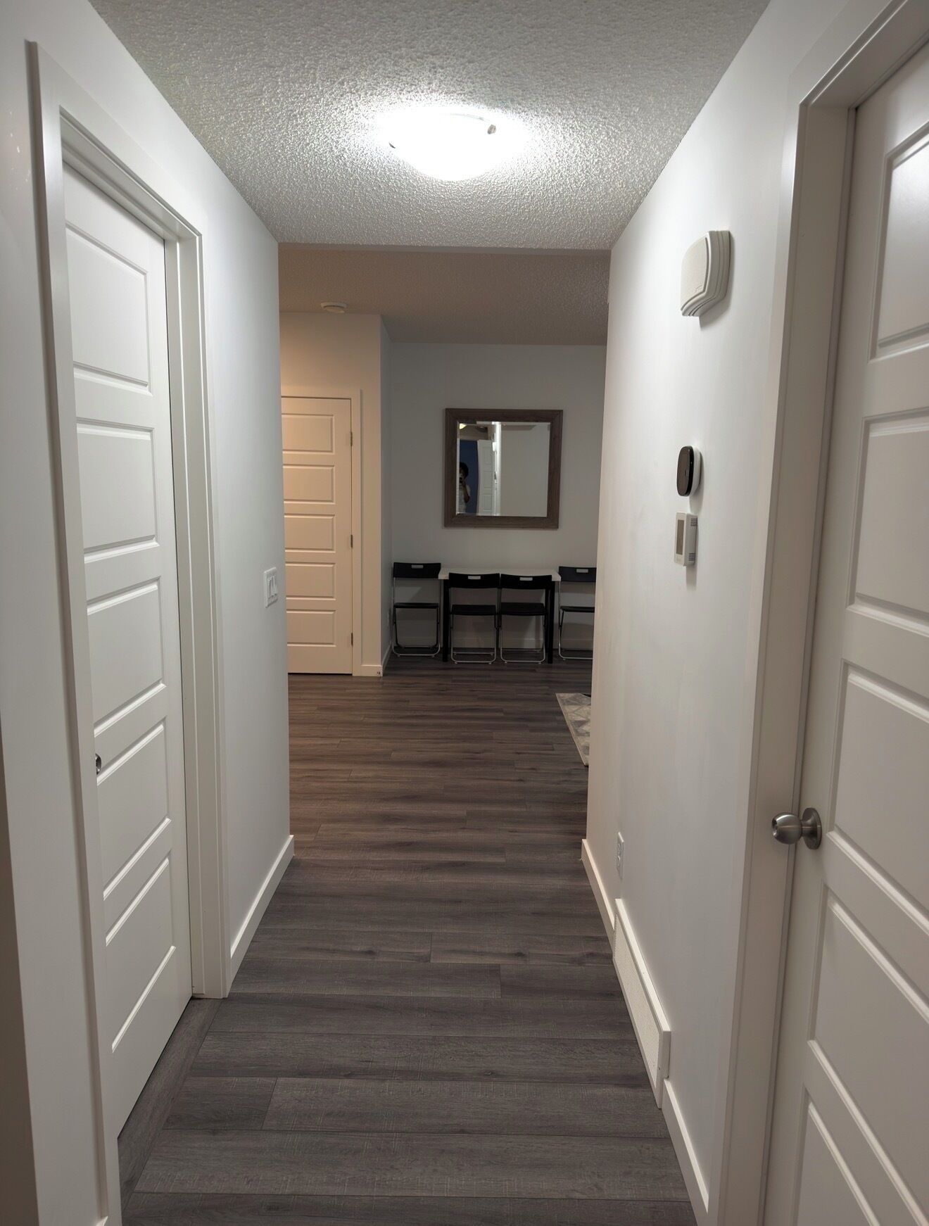 A hallway with a wooden floor, white walls, and a ceiling light. There are doors on both sides, and a dining area with chairs and a table is visible in the background.