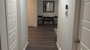 A hallway with a wooden floor, white walls, and a ceiling light. There are doors on both sides, and a dining area with chairs and a table is visible in the background.