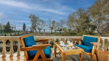 A terrace with two wooden chairs and a table, a glass-top coffee table with cups and a plate of food, and a view of trees and a distant building.