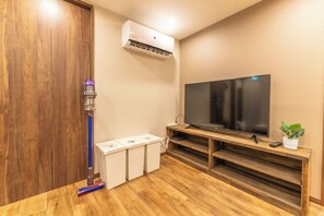 Ein Zimmer mit Holzboden, einem an der Wand montierten Fernseher auf einem Holzständer und einer Wand-Klimaanlage. Ein Dyson Staubsauger ist ebenfalls vorhanden.