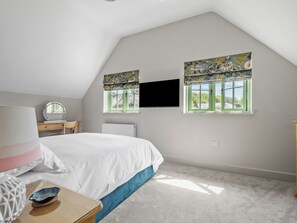 Une chambre à coucher comprenant un lit, un bureau, une chaise, un téléviseur et deux fenêtres avec des rideaux.
