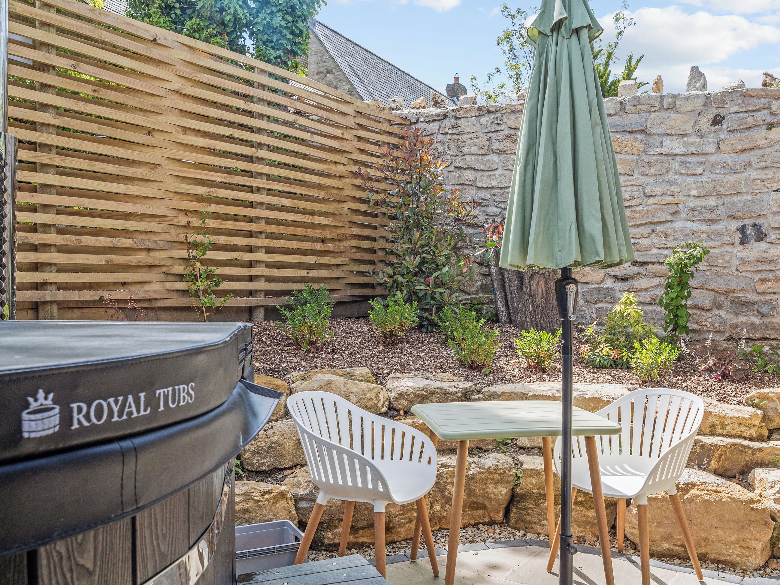 A barbecue grill labeled 'ROYAL TUBS,' white plastic chairs, a small table, and a stone wall with plants.