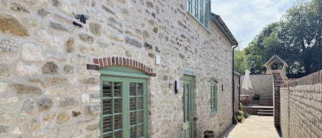 Un edificio de piedra con un estrecho sendero, una ventana pequeña con contraventanas y un muro de ladrillo.