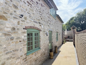 A stone building with a narrow pathway, a small window with shutters, and a brick wall. - Suncrest Cottage - UKC9942 (Weymouth)
