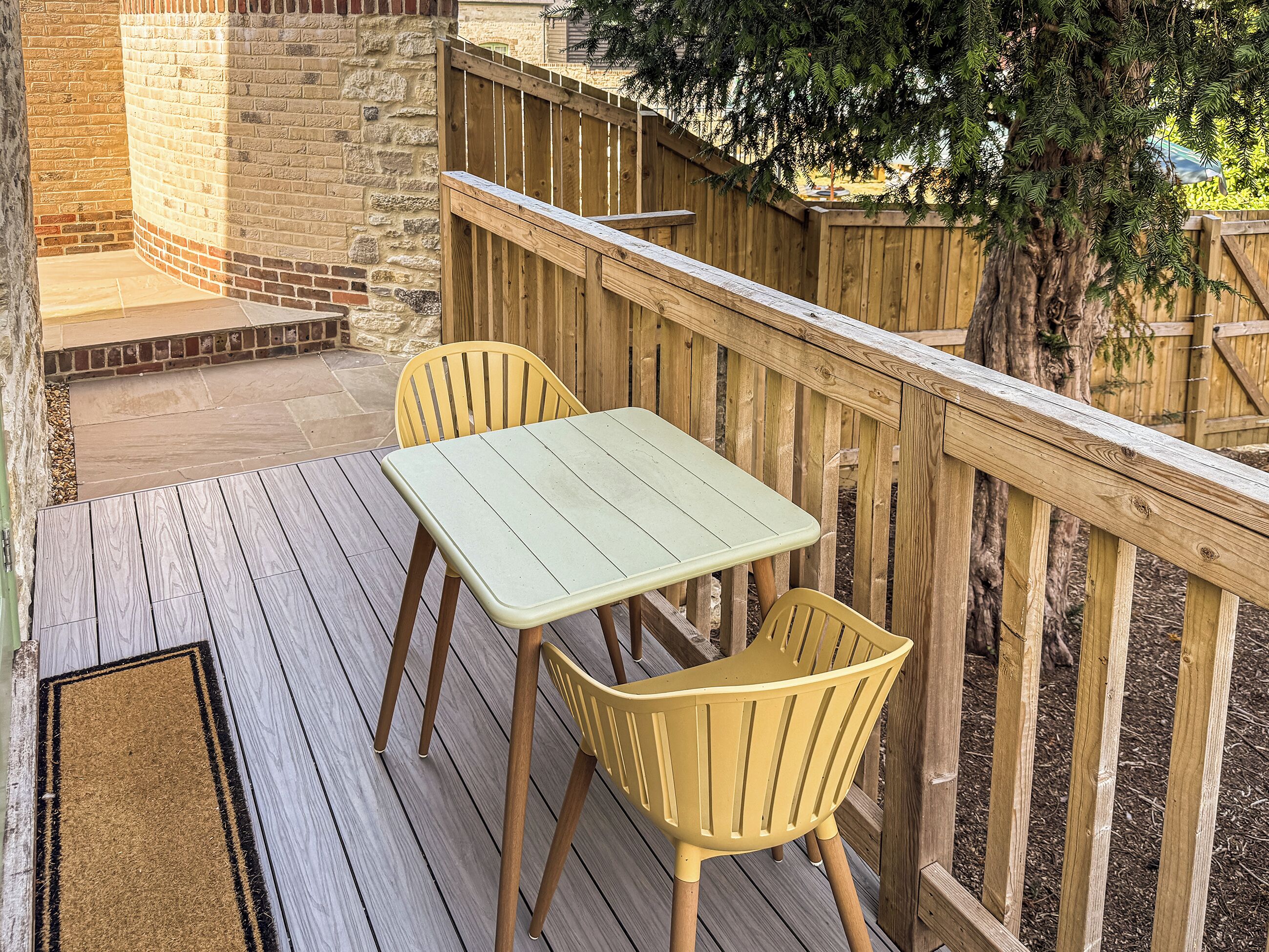Una terraza de madera con una mesa y dos sillas, rodeada por una valla de madera y un muro de ladrillo.