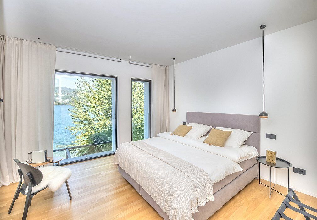 Une chambre à coucher moderne avec un grand lit, une chaise, une petite table et une vue sur l’extérieur.