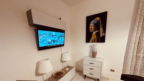 Un salon moderne avec un téléviseur à écran plat fixé au mur, une table d’appoint avec une lampe et des objets décoratifs, un meuble blanc avec des tiroirs, et un tableau représentant une femme portant un foulard bleu et jaune.