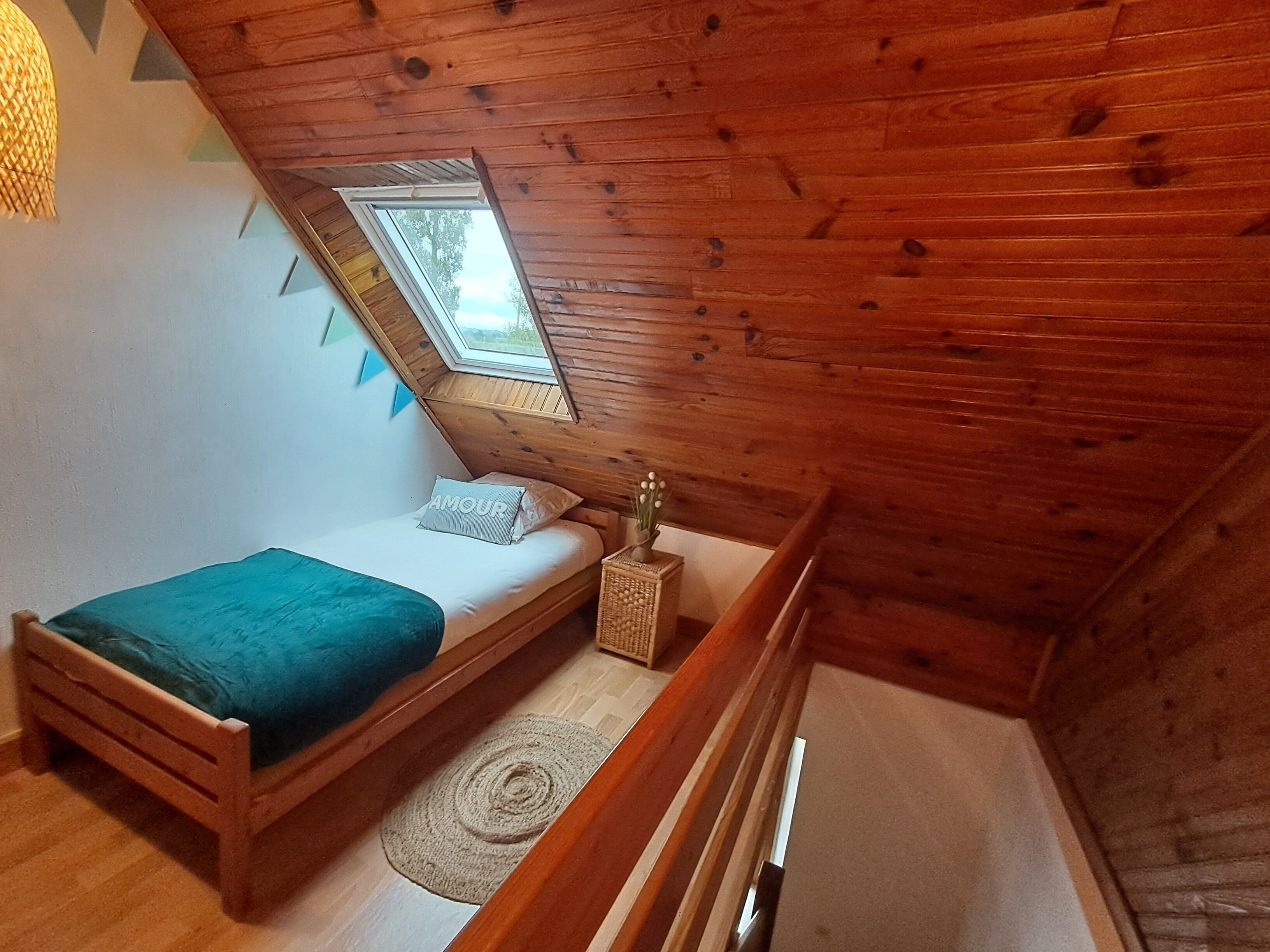 A small, cozy room with a sloped ceiling, a single bed with a blue blanket, a small table with a vase, and a skylight.