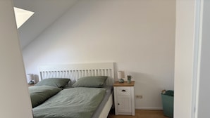 A bedroom with a sloped ceiling, a bed with green bedding, a bedside table with a lamp, a small cabinet, and a white rug on the wooden floor.