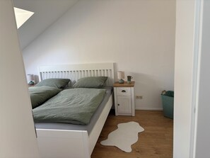 A bedroom with a sloped ceiling, a bed with green bedding, a bedside table with a lamp, a small cabinet, and a white rug on the wooden floor.