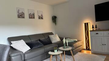 Ein modernes Wohnzimmer mit einem grauen Ledersofa, zwei kleinen Beistelltischen, einem weißen Teppich und einem Schrank mit einem Fernseher darüber.