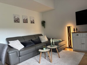 A modern living room with a grey leather sofa, two small side tables, a white rug, and a cabinet with a TV above.