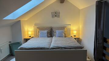Ein Schlafzimmer mit schrägem Dach, einem Oberlicht, einem Bett mit zwei Kissen, zwei Nachttischlampen und einem kleinen Wandbild.